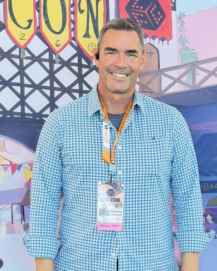 CJ Connoy smiling at LFG Con 2026, wearing his Special Guest badge in front of the con's illustrated tabletop backdrop